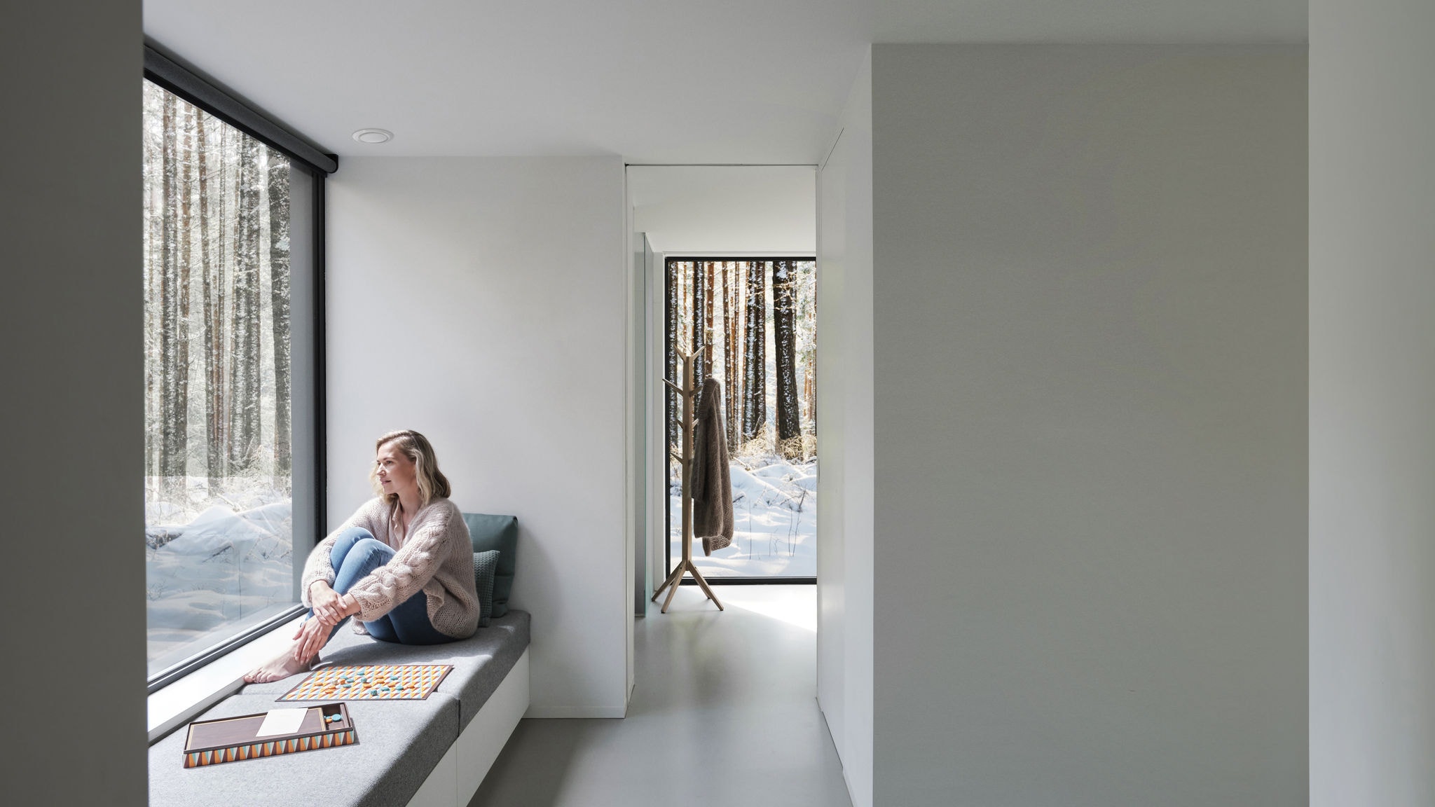 Širokouhlá fotka bosej ženy, ktorá sedí pred oknom a pozerá sa von na stromy a sneh.