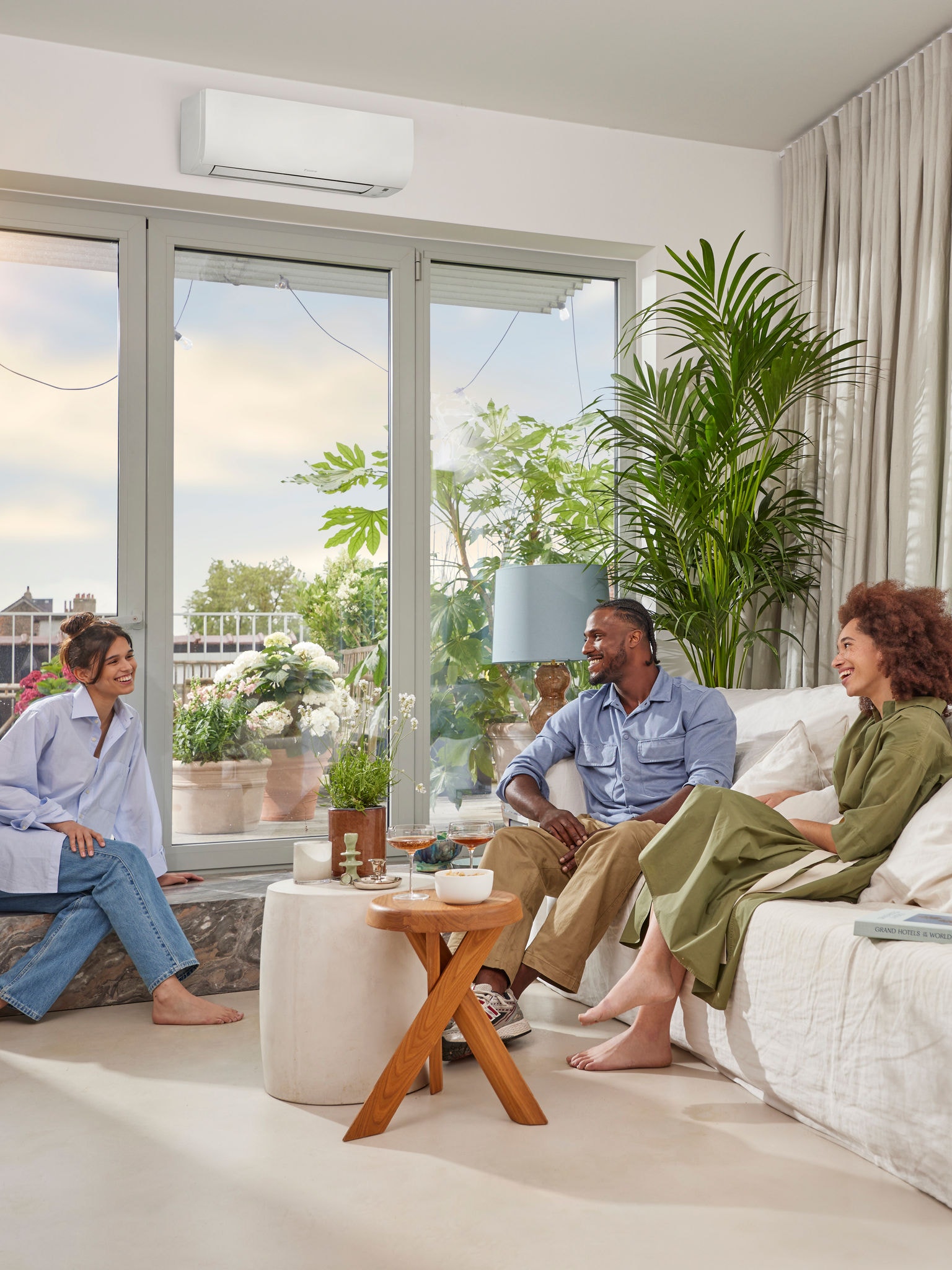 Muž a dve ženy sedia a rozprávajú sa počas slnečného dňa vo svetlej, vzdušnej miestnosti s bielymi stenami a nenápadnou bielou nástennou klimatizáciou nainštalovanou nad veľkým oknom.