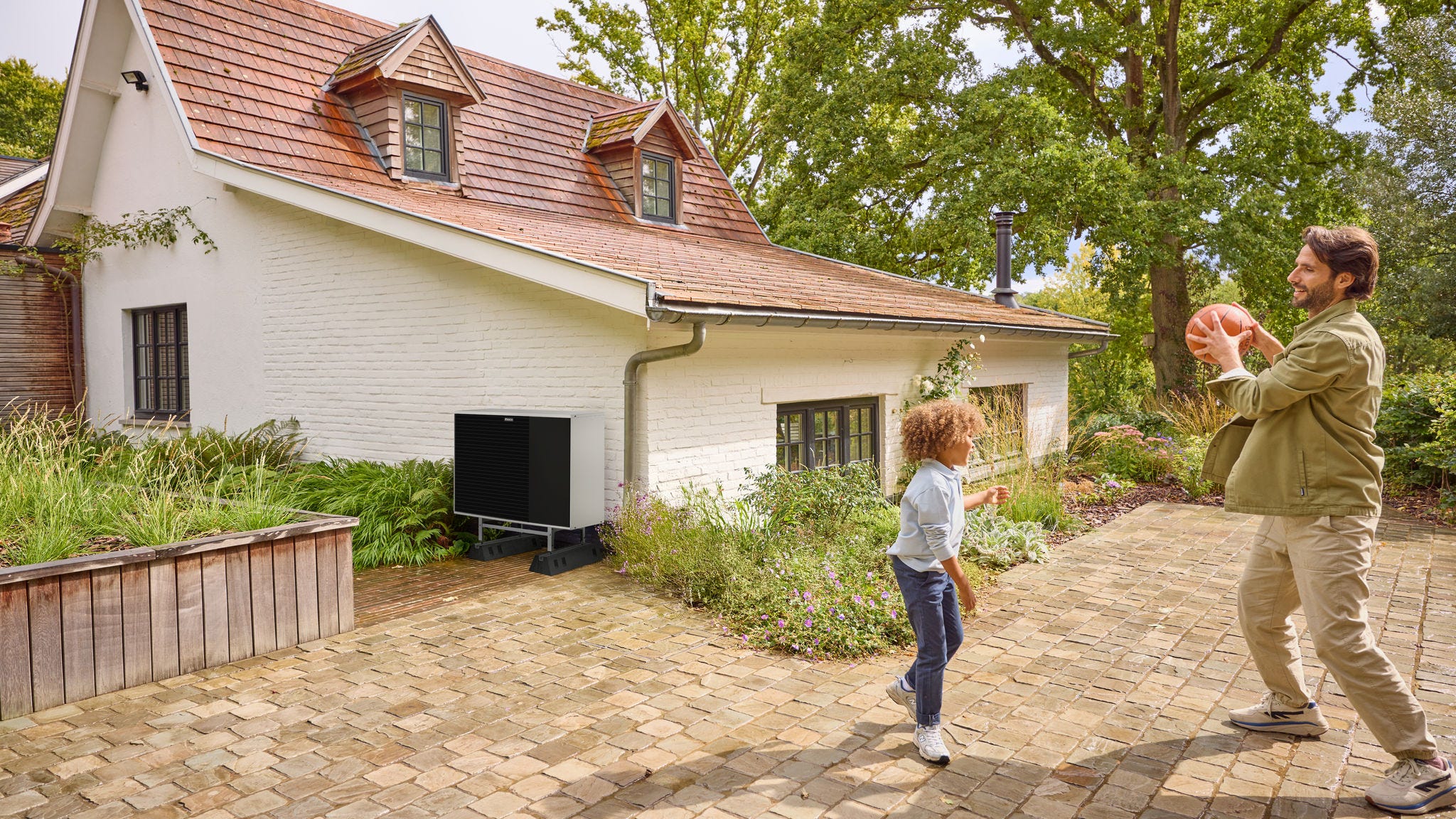 Otec s dieťaťom sa hrajú s loptou v záhrade, kde je nainštalované tepelné čerpadlo.