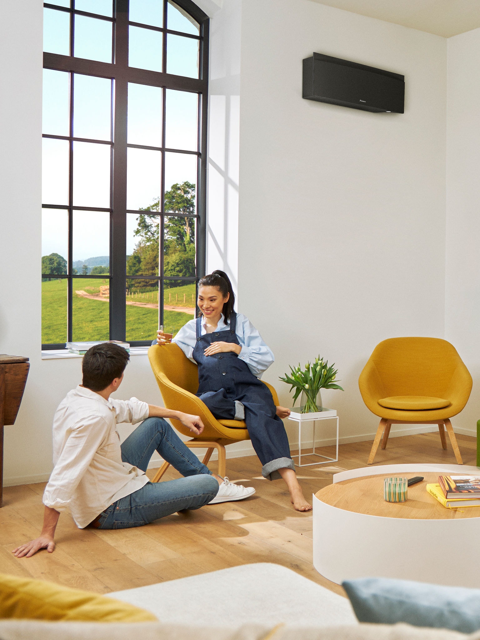 Muž a žena sedia pred veľkým oknom v svetlej miestnosti s čiernym nástenným tepelným čerpadlom na stene za nimi