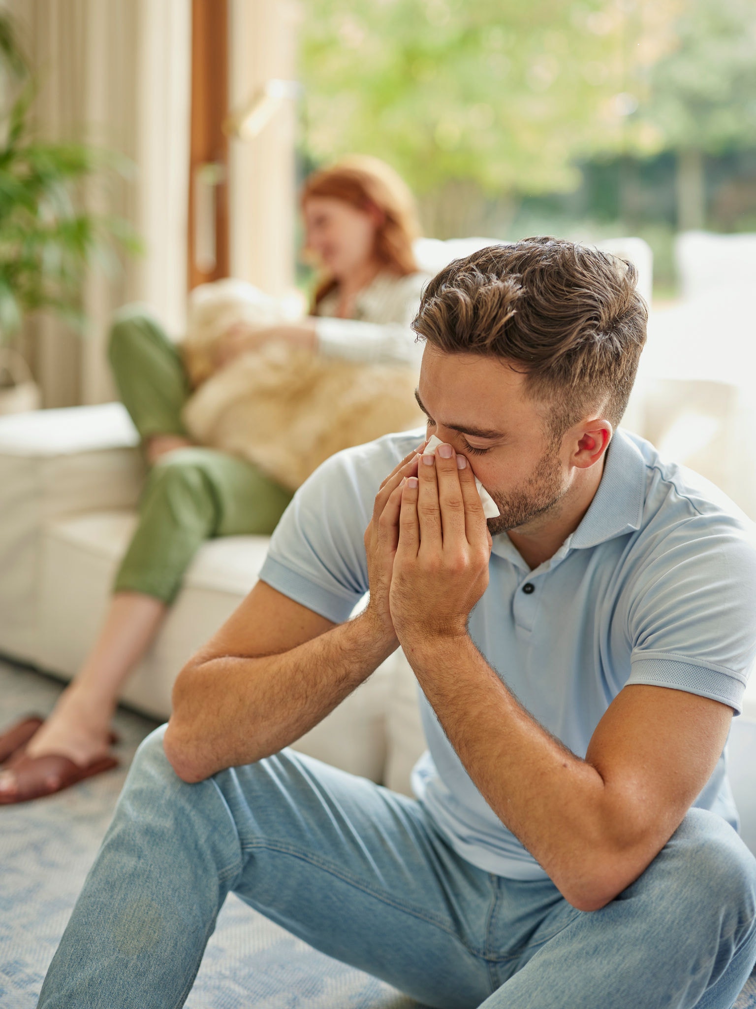 Žena v pozadí sedí na mäkkých prikrývkach a v lone má zlatého retrievera, zatiaľ čo muž v popredí kýcha.