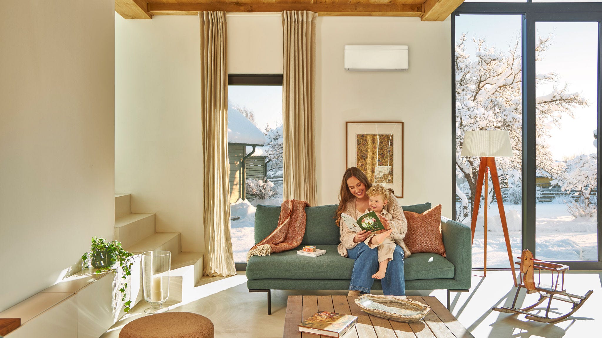 Matka sedí v teple na gauči pod nástenným tepelným čerpadlom vzduch-vzduch a svojmu dieťaťu sediacemu na kolenách číta knihu, zatiaľ čo vonku sneží